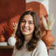 Portrait of a smiling young woman, mid-20s, with a warm, inviting expression, against a soft terracotta and white background.